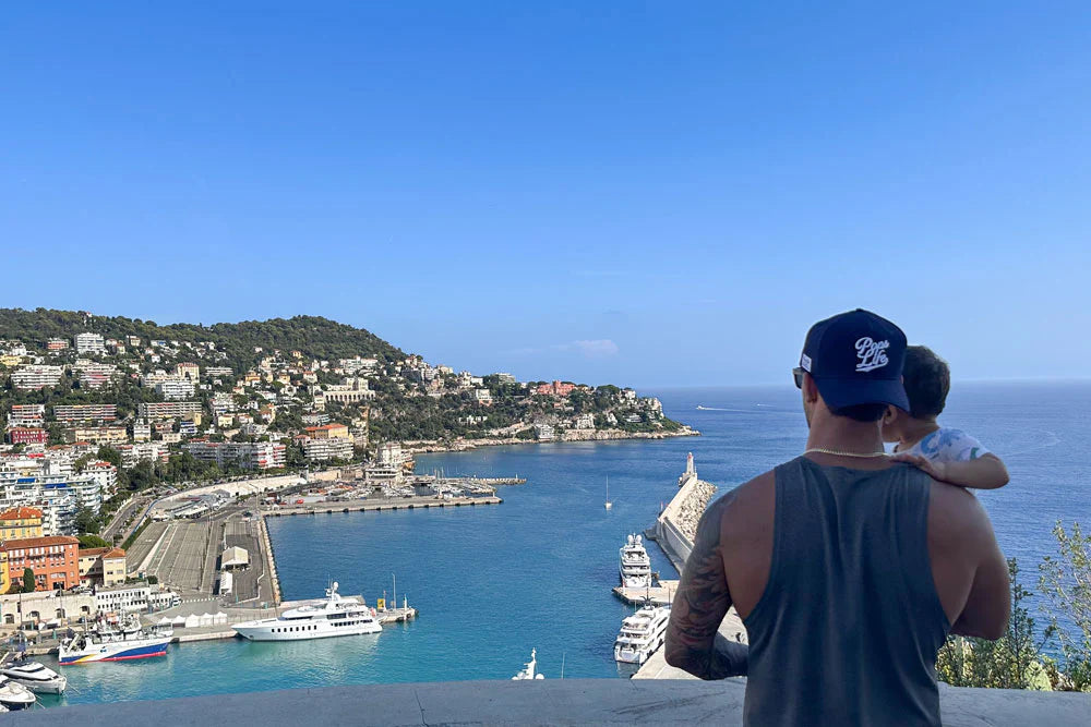 A father wearing a navy “Pops Life” dad hat holds his child while overlooking the vibrant harbor and hillside of Nice, France, highlighting the effortless style and versatility of dad hats.
