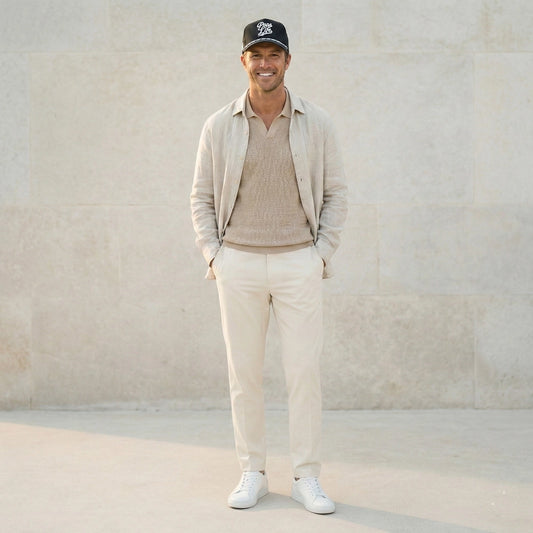 A man wearing a Pops Life Black Dad Hat with a white rope, a beige sweater, pants, and a cap, standing against a light stone wall.
