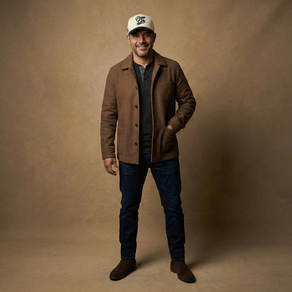 A man wearing a brown coat, a white cap, and dark jeans and a Pops Life Tan and Black Dad Hat against a beige background