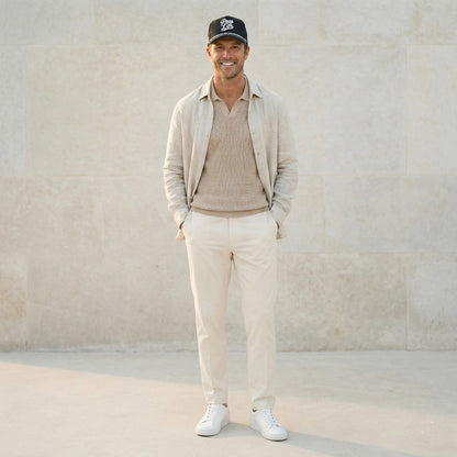 A man wearing a Pops Life Black Dad Hat with a white rope, a beige sweater, pants, and a cap, standing against a light stone wall.