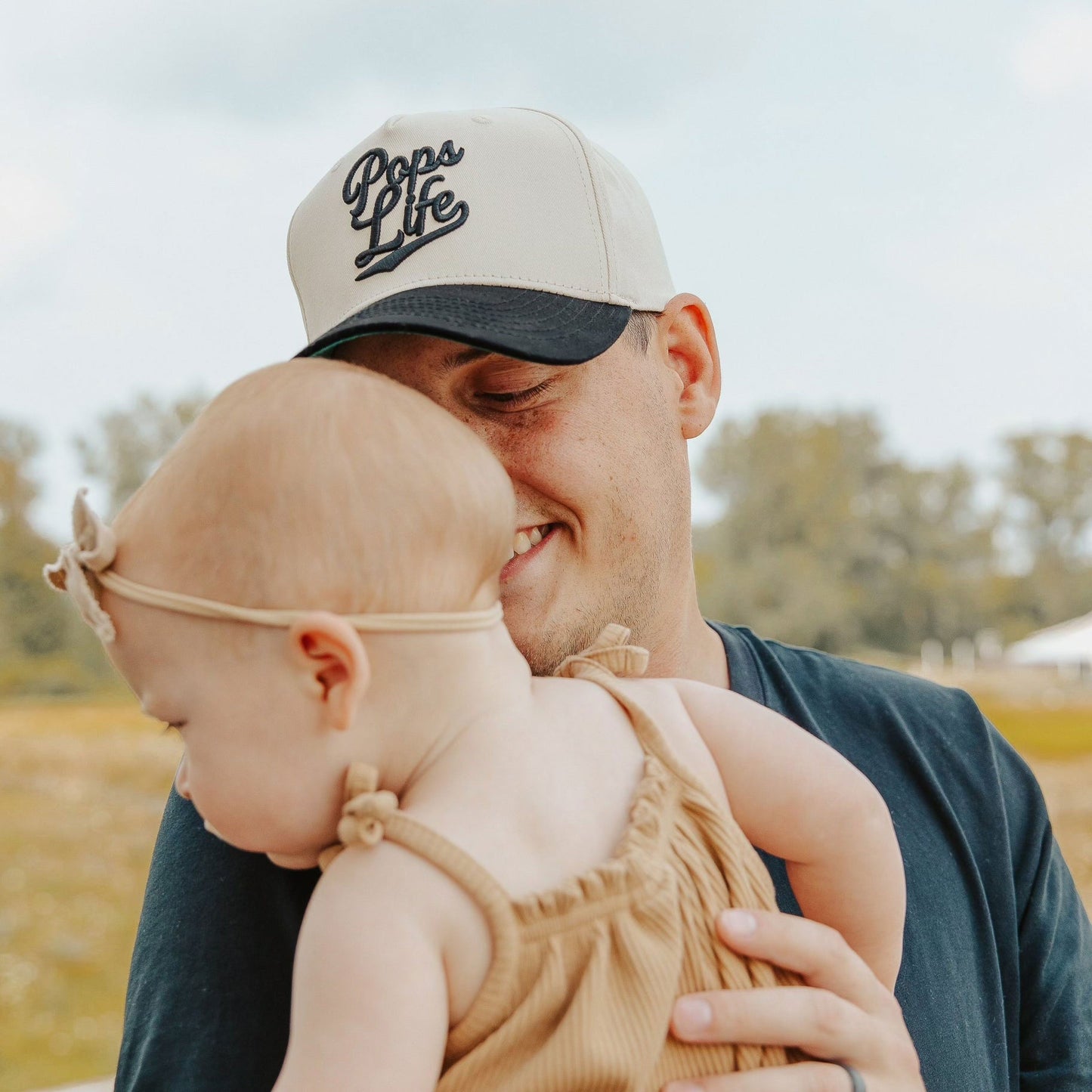 Dad wearing Pops Life A-Frame hat, holding his baby close outdoors.