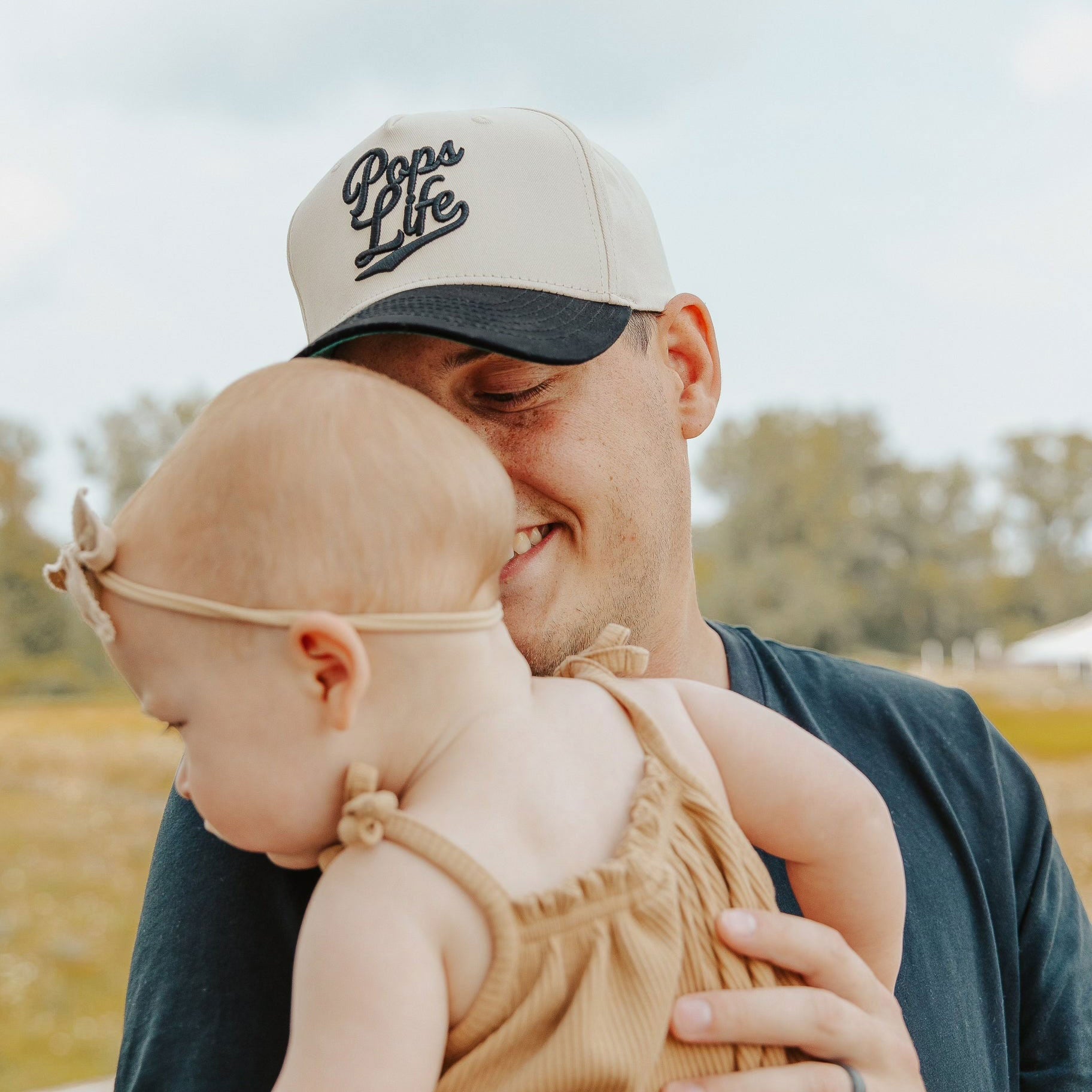 Dad wearing Pops Life A-Frame hat, holding his baby close outdoors.