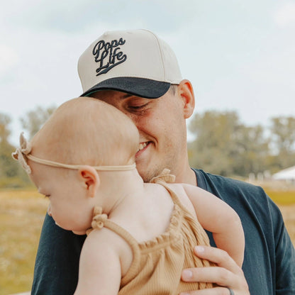 Man wearing a Pops Life Tan and Black Dad Hat  with 'Pops Life' text, holding a baby in an outdoor setting.