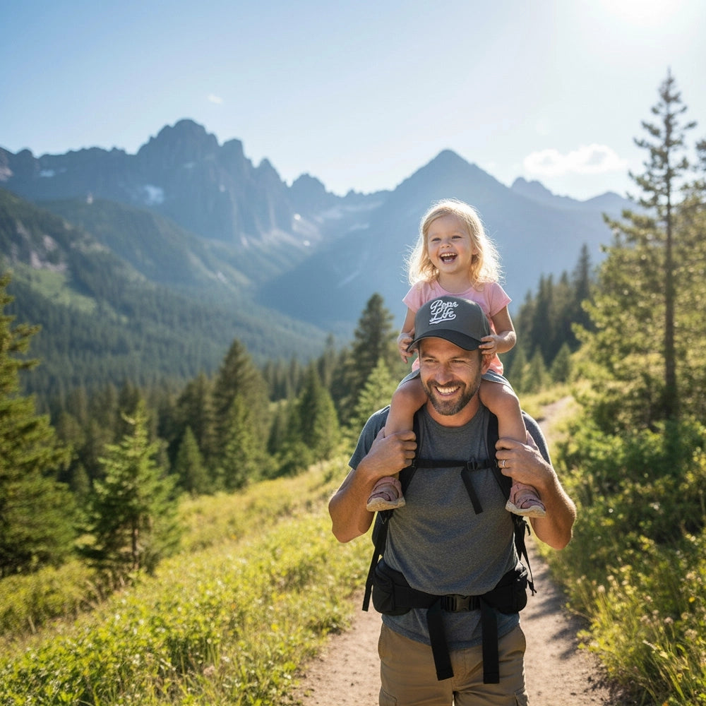 Man wearung a Pops Life Gray and Black Dad Hat carrying a child on his shoulders with mountains and forest in the background