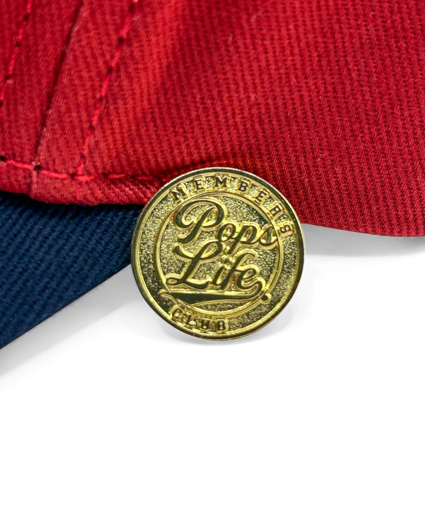 A close-up of the exclusive Pops Life member pin in gold, featuring intricate detailing and the Pops Life logo, placed against the maroon and navy fabric of the hat.