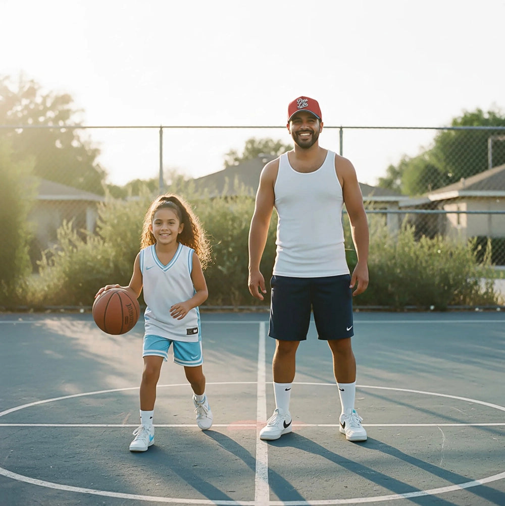 A dad wearing a Pops Life Maroon Blue Dad Hat stands on an outdoor basketball court with his daughter, who is dribbling a basketball. 