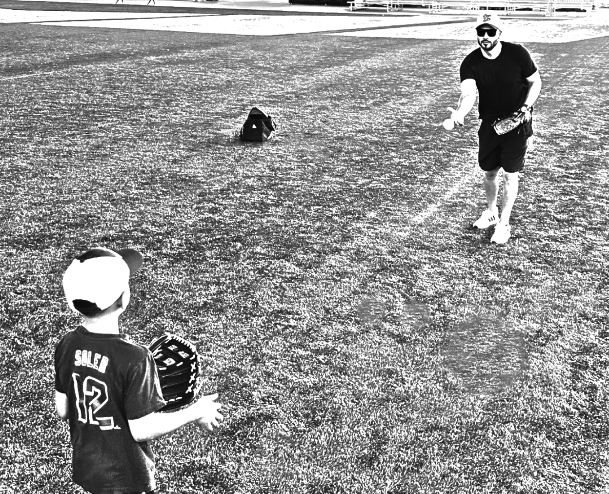 Black and white image of a dad playing catch with his child, both wearing best dad hats.